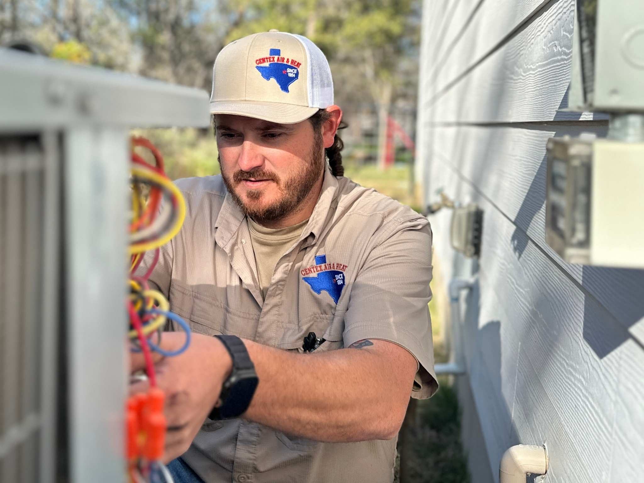 Technician inspecting low-voltage electrical wiring on an outdoor AC condenser unit against a blue sky in Bastrop County